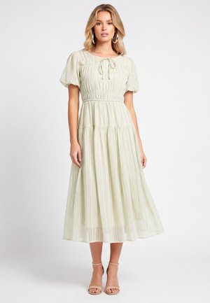 Femme portant une robe midi vert clair avec des manches bouffantes, un col noué et des talons beiges à bride à la cheville sur un fond uni.
