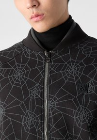 Svart zip-up jakke med et edderkoppnett mønster i grått. Har en ribbet krage og en logo-patch på brystet, og har en glatt tekstur.