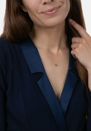 Goldene Halskette mit einem runden Anhänger, getragen über einem tiefblauen Blazer mit satiniertem Kragen, der eine glatte Textur und dezenten Glanz aufweist.