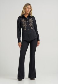 Camisa preta de botão com mangas longas, apresentando um intrincado bordado dourado na frente. Combinada com calças pretas de corte amplo e sapatos pretos brilhantes.