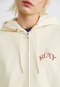 Kremfarget hettegenser med glidelås, snor og en lodden innerside. Har "ROXY" trykt i brunt med et solmotiv.