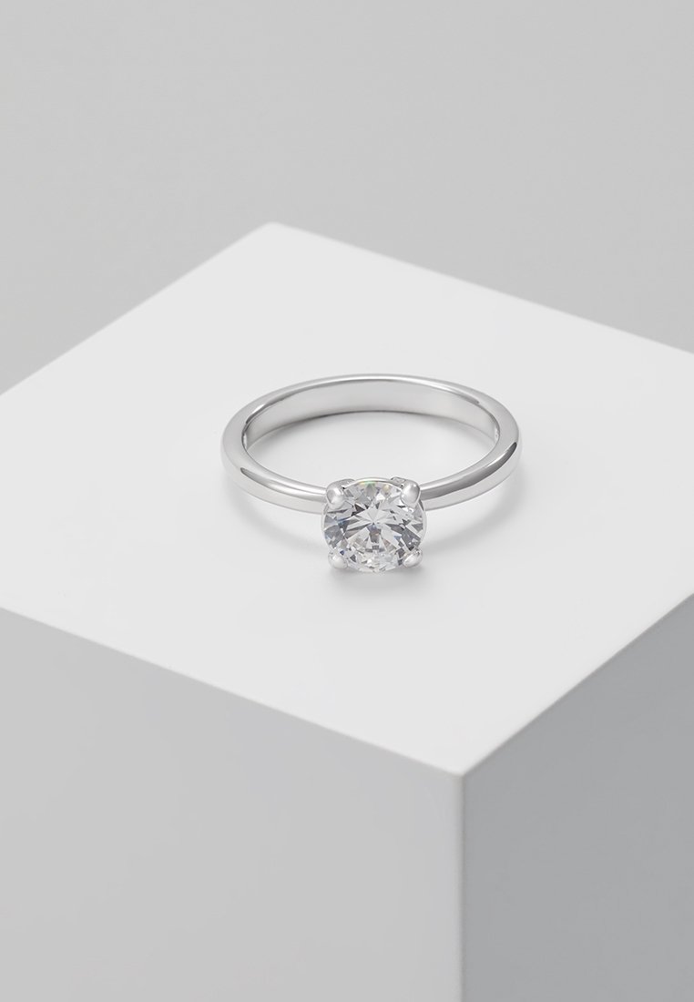 Bague solitaire en argent avec une pierre précieuse ronde et claire sertie de quatre griffes, posée sur une surface géométrique blanche.