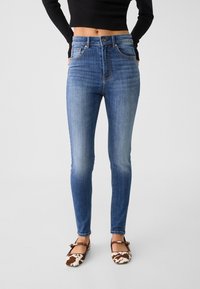 Höga blå denimjeans med en figurnära silhuett, subtila slitningar och kontrasterande sömmar, ihop med bruna och vita mönstrade skor.