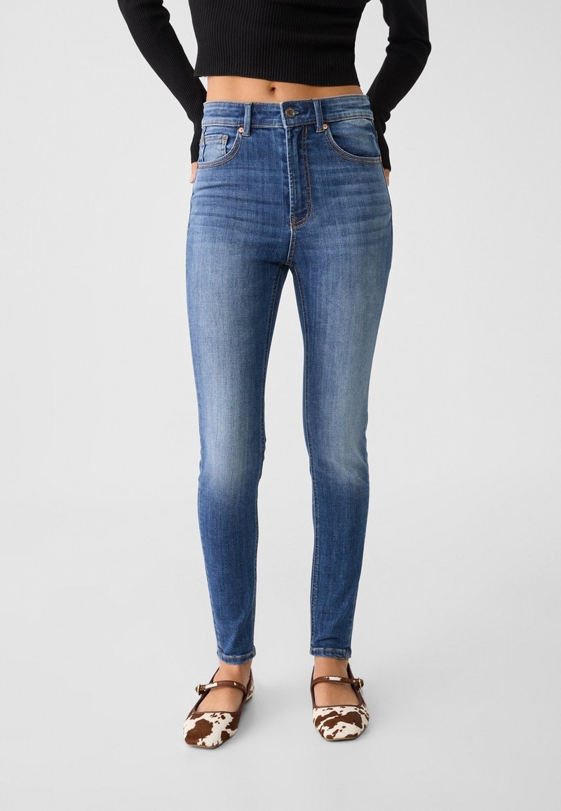 Höga blå denimjeans med en figurnära silhuett, subtila slitningar och kontrasterande sömmar, ihop med bruna och vita mönstrade skor.