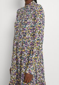 Blommig klänning med hög halsringning, långa ärmar och rynkad textur. Har ett livfullt mönster av lila, gula och vita blommor p�å en mörk bakgrund.