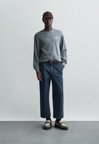 Pilkas ilgas rankovės džemperis su logotipu, mėlyni plačios kojų kelnės, ruda kepurė ir daugiaspalviai batai su dirželiais ir sagtimis. Vienalytės tekstūros.