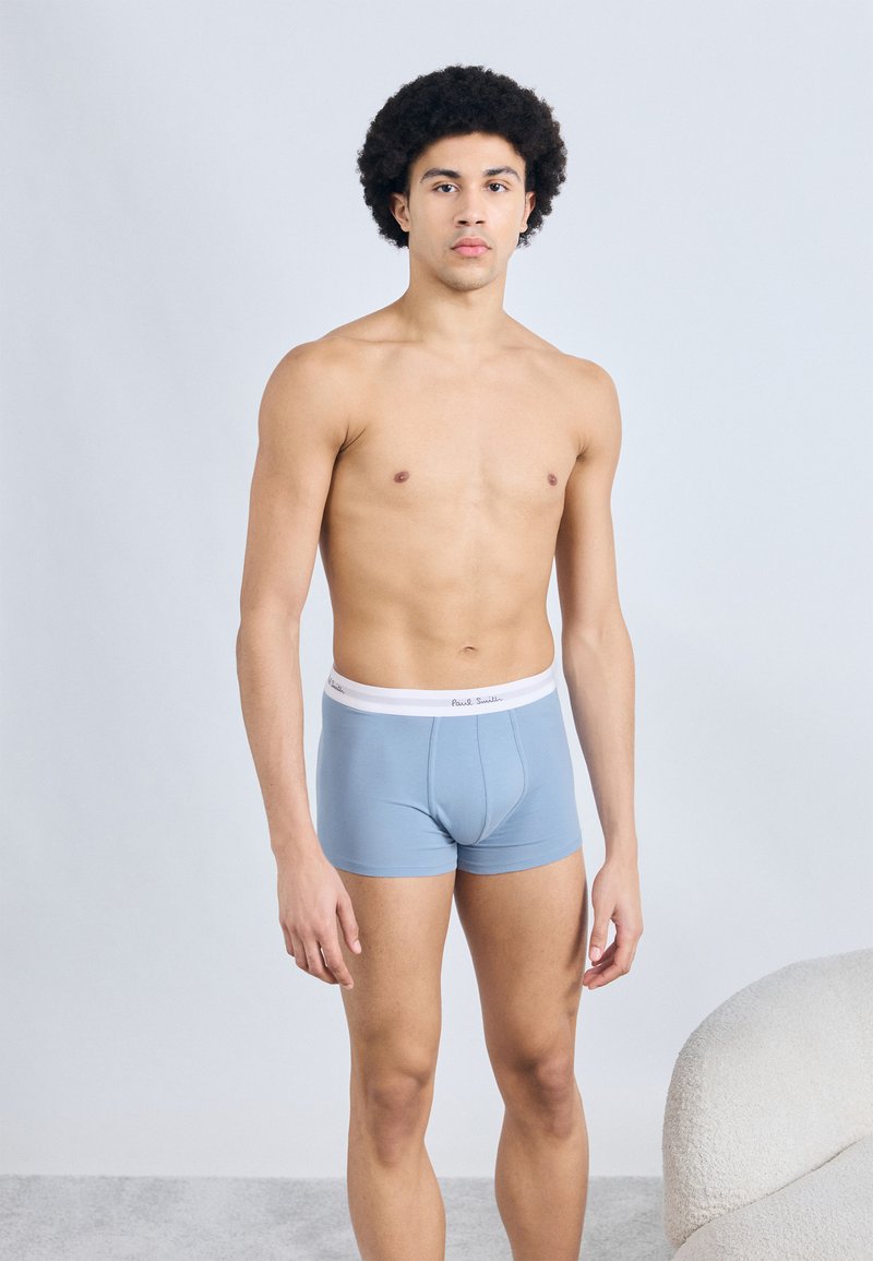 Caleçons courts bleu clair avec une ceinture blanche, arborant un logo, portés par un mannequin se tenant devant un fond uni.