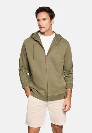 Hombre con sudadera con cremallera de color verde oliva y pantalones cortos beige, de pie con las manos en los bolsillos contra un fondo liso.