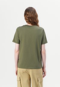 Osoba z długimi brązowymi włosami, widoczna z tyłu, ubrana w oliwkową koszulkę z krótkim rękawem i beżowe spodnie cargo na jednolitym tle.