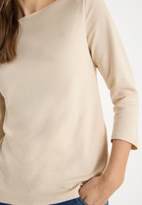 Beige, long-sleeve top made of soft fabric, featuring a wide neckline and a relaxed fit, paired with denim. The texture appears smooth.