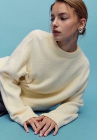 Cremefarbener Pullover aus weicher Wolle mit einem gerippten Kragen und Bündchen. Das Modell liegt auf blauem Hintergrund und hält die Hände vor sich verschränkt.