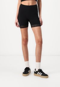Black shorts featuring a lace trim at the hem, made from a smooth material. They have a fitted design and are paired with black sneakers.
