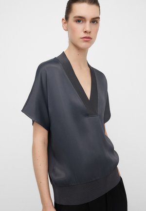 Junge Frau trägt eine lockere, dunkelgraue Satinbluse mit tiefem V-Ausschnitt und kurzen Ärmeln, steht vor einem schlichten weißen Hintergrund.