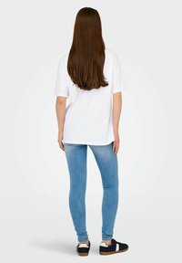 Bílý oversize tričko, světle modré skinny džíny, černé tenisky s bílými akcenty. Dlouhé hnědé vlasy, pohled zezadu.