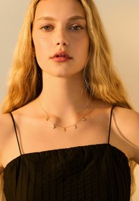 Collier en or avec cinq pendentifs géométriques, porté sur une robe noire texturée à fines bretelles, présenté sur un fond doucement éclairé.