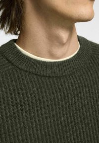 Suéter de canalé verde oscuro con una superficie texturizada, que presenta un escote redondo sobre una camisa de color claro.