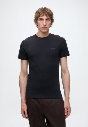 Giovane uomo con capelli ricci castano chiaro che indossa una T-shirt nera aderente a maniche corte e pantaloni marrone scuro, in piedi davanti a uno sfondo chiaro e semplice.