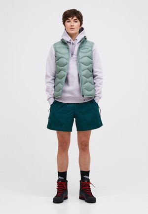 Person trägt eine hellgrüne gesteppte Weste über einem lavendelfarbenen Kapuzenpullover, tealfarbene Shorts, schwarze Socken und schwarze Wanderschuhe mit roten Schnürsenkeln, steht.