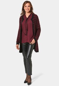 Burgunderfarbener und schwarzer gemusterter Strickcardigan über einer weinroten Bluse mit Schleife, kombiniert mit glänzenden schwarzen Hosen und schwarzen High Heels.