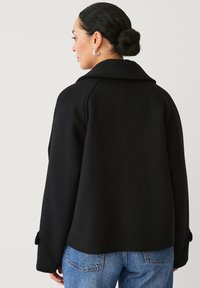 Schwarzer Woll-Mix-Mantel mit breitem, eingekerbtem Kragen, lässiger Passform und Knopfdetails an den Ärmeln; kombiniert mit hellblauen Jeans.
