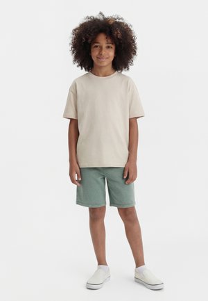 Παιδί με σγουρά μαλλιά στέκεται χαμογελώντας, φορώντας μπεζ κοντομάνικο πουκάμισο, πράσινα σορτς, άσπρες κάλτσες και λευκά παπούτσια χωρίς κορδόνια.