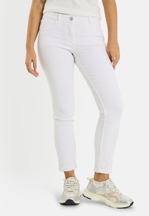 Mujer con vaqueros blancos de corte ajustado, remangados en los tobillos, y zapatillas blancas con detalles en beige y plata, de pie contra un fondo blanco.