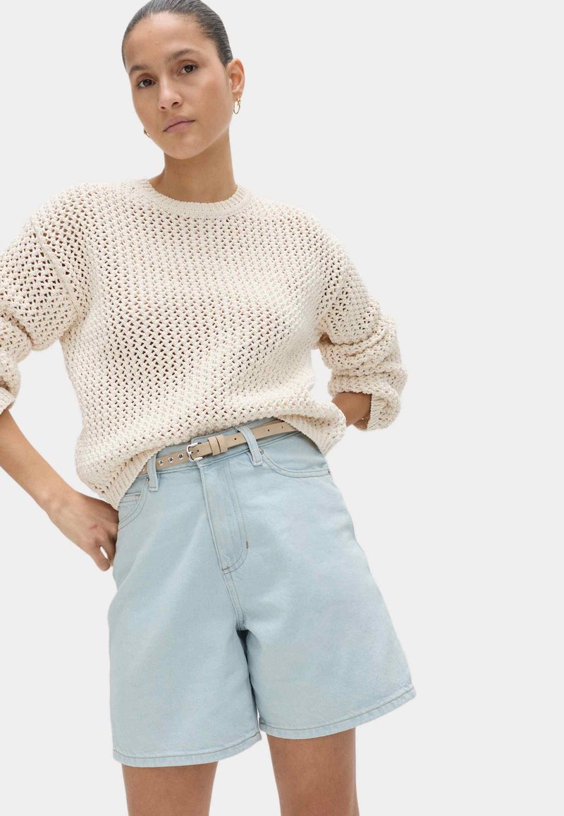 Mujer con un suéter de punto color crema con mangas remangadas y shorts de mezclilla de cintura alta color azul claro con un cinturón beige, de pie con las manos en la cadera.
