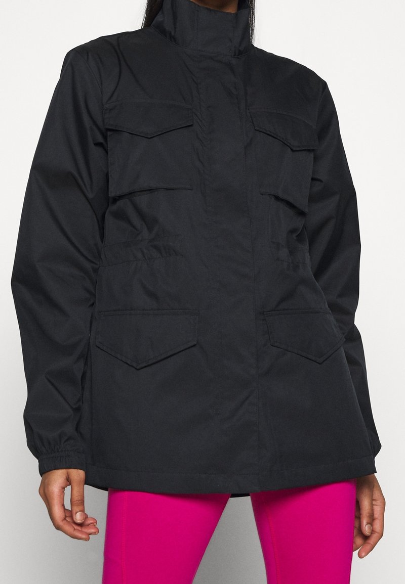 Svart lättviktsjacka med hög krage, flera framfickor och figursydd passform, kombinerad med ljusrosa leggings.