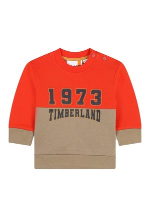 Maglione rosso e beige con maniche lunghe, scollo rotondo e dettagli con bottoni sulla spalla. Presenta la scritta "1973 TIMBERLAND" in lettere scure.