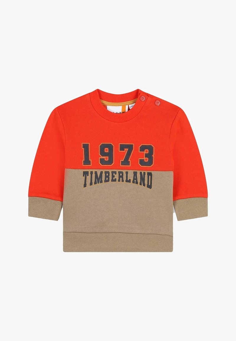 Maglione rosso e beige con maniche lunghe, scollo rotondo e dettagli con bottoni sulla spalla. Presenta la scritta "1973 TIMBERLAND" in lettere scure.
