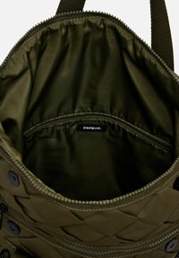 Intérieur d'un sac matelassé vert olive avec doublure lisse, comportant une poche zippée et une étiquette. L'ouverture a un bord cousu et des boutons.