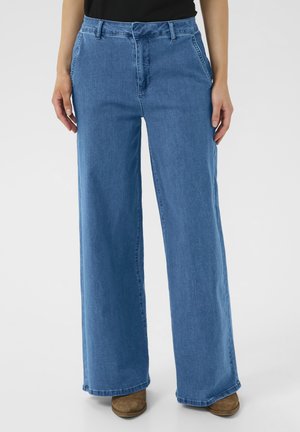 Pantalons en denim bleu à jambe large avec une taille haute, présentant des poches avant et une texture lisse, associés à des bottes à talons en daim marron.