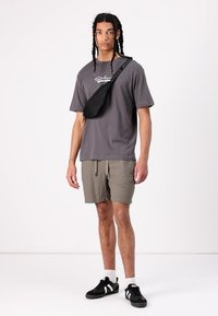 Camiseta gris con un logo, pantalones cortos a cuadros claros, riñonera negra y zapatillas negras con acentos blancos. Fondo liso.