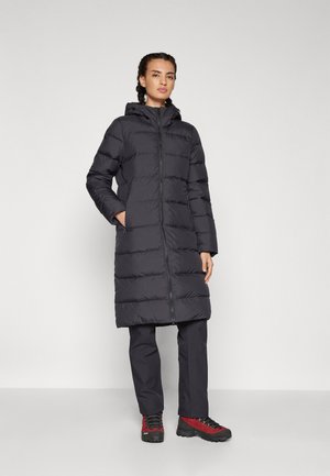Giacca nera imbottita con cappuccio, chiusura con zip e tasche laterali. Tessuto liscio con motivo a righe orizzontali. Indossata con pantaloni scuri e scarpe rosse.