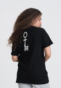Sort bomulds t-shirt med et sølvfarvet grafisk print af et kønssymbol og et ansigt på ryggen. Kortærmet og afslappet pasform.