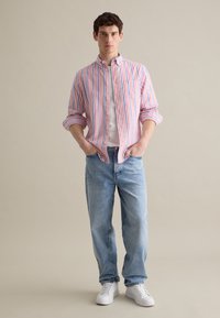 Camisa de botones a rayas en rosa, azul y blanco; combinada con jeans de color azul claro y zapatillas blancas. La camisa cuenta con un bolsillo en el pecho y mangas enrolladas.