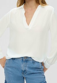 Blusa bianca leggera con scollatura a V, maniche lunghe e una texture liscia, abbinata a pantaloni corti di denim blu chiaro.