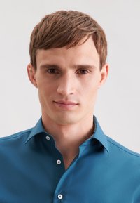 Junger Mann mit kurzem braunem Haar, der ein blaues Button-Up-Hemd trägt, schaut mit einem neutralen Ausdruck vor sich hin vor einem schlichten, hellen Hintergrund.