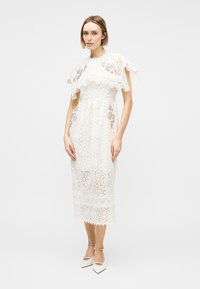 Naine, kes kannab valget pitsist midi-kleiti lõhustatud kapuutsiga ja valgeid terava ninaga kontsakingi, millel on pahkluu rihmad.