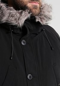 Parka noire avec col en fausse fourrure, dotée d'une fermeture à boutons, d'une capuche avec cordon de serrage et d'un tissu texturé. Des poches sont visibles à l'avant.