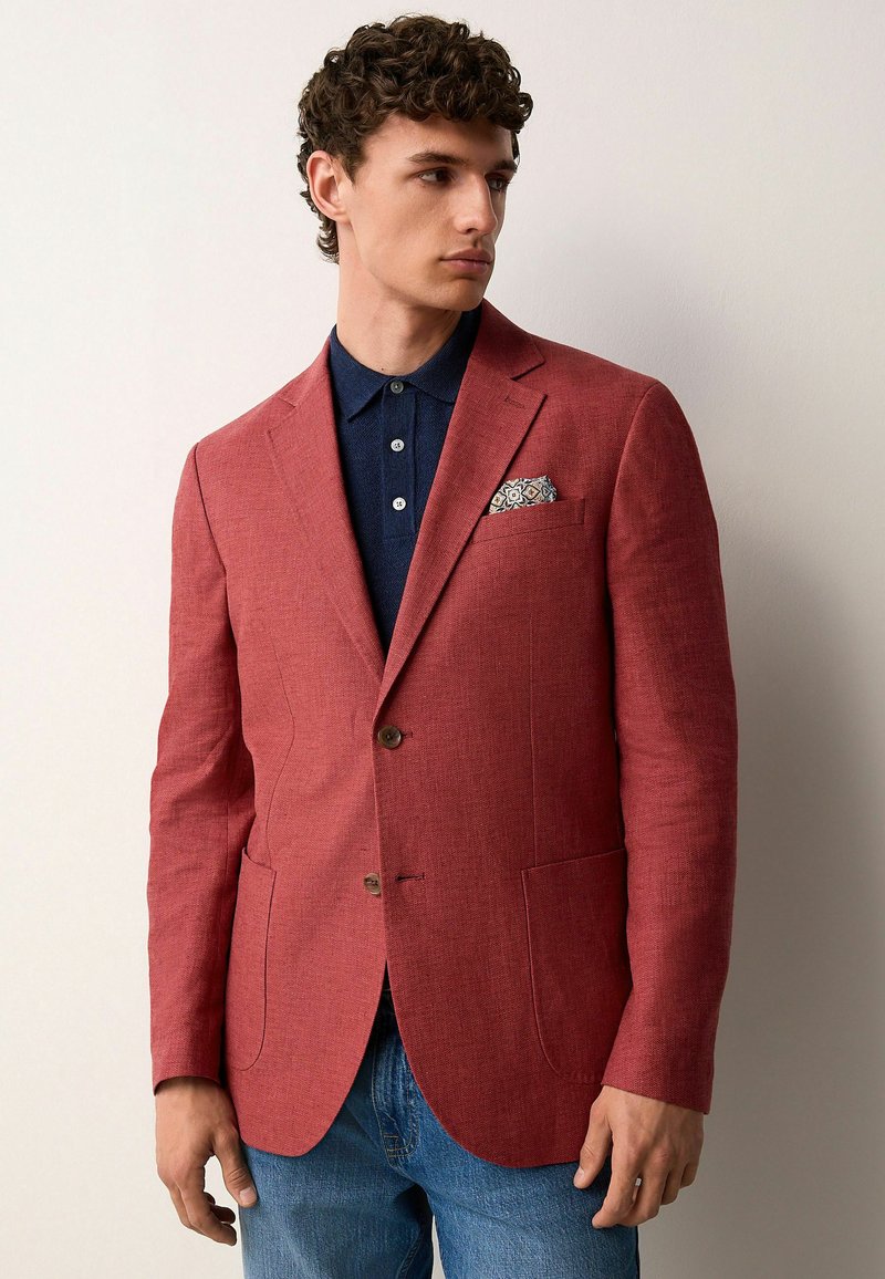 Roter Leinenblazer mit zwei Knöpfen, Reverskragen und Taschentaschen. Kombiniert mit einem dunkelblauen Poloshirt und einem gemusterten Einstecktuch.