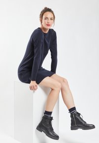 Marineblauwe gebreide jurk met lange mouwen, ontspannen pasvorm en lengte boven de knie. Gepaard met zwarte veterschoenen tot de enkel en gestreepte sokken.