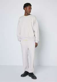 Siv svečanik večje velikosti in usklajene trenerke, narejene iz mehkega blaga, z okroglem izrezom in rebrastimi manšeti, obute z črnimi čevlji.