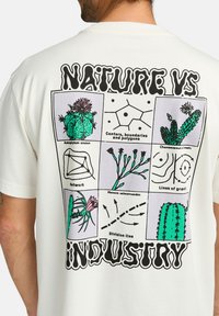 Cream-colored t-shirt featuring a graphic design with cacti illustrations, black outlines, and text reading "NATURE VS INDUSTRY."