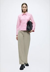 Ροζ ριγέ πουκάμισο με κουμπιά, μπεζ παντελόνι φαρδύ, και μαύρη τσάντα. Το μοντέλο φοράει chunky μαύρα παπούτσια, στέκεται μπροστά σε ένα ουδέτερο φόντο.