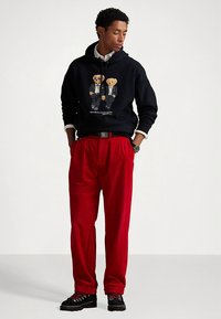 Svart hettegenser med bjørnegrafikk, røde plisserte bukser og svarte joggesko. Hettegenseren har snøring og en avslappet passform.