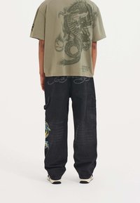 Olive green oversized t-shirt with a dragon graphic, paired with black cargo pants featuring colorful embroidery and side pockets.