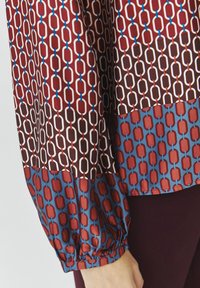 Langmouw blouse met een kettingpatroon in rood, bruin en blauw, met een geplooide mouwmanchet en een contrasterend onderkant.