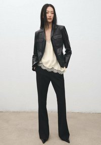 Chaqueta de cuero negra con dos bolsillos en el pecho, sobre una blusa crema con encaje, combinada con pantalones negros de talle alto y zapatos puntiagudos.