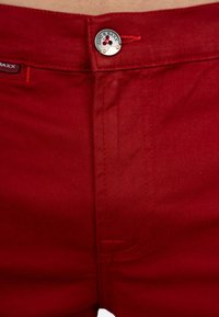 Pantaloni in cotone rosso misto con chiusura a bottone in metallo con branding, cuciture visibili e una texture liscia.